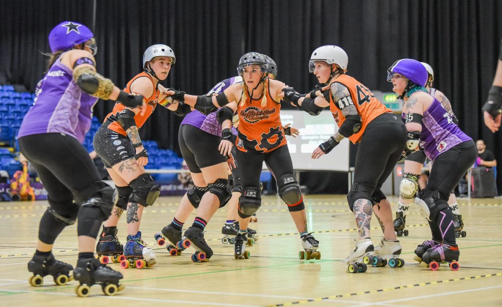 Three Sheffield Steel skaters dressed in orange face of gainst a jammer dressed in purple