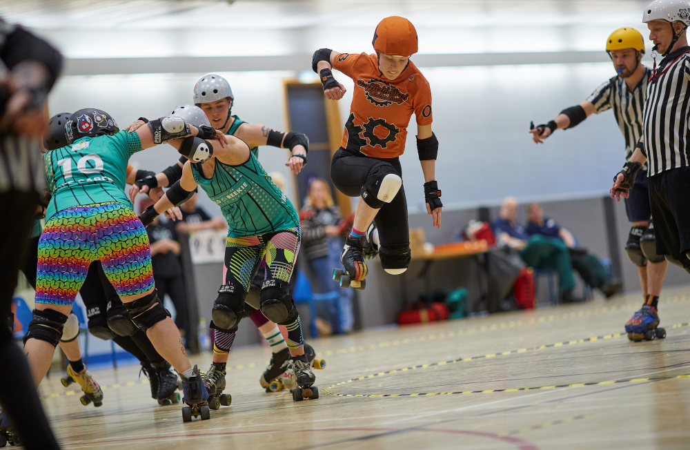 An SSRD skater, Wince, is leaping into the air in order to clear the apex of the track and avoid the three opposing skaters looking to hit Wince off track