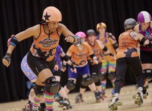A roller derby jammer wearing an orange jammer star and an orange Sheffield steel roller derby runiform skates out of pack whilst her team mates block the opposing team wearing a purple uniform